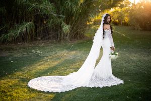 Bride in elegant white lace gown with long train and veil holding bouquet on green lawn with trees.