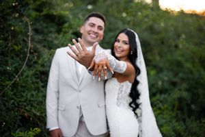 Newlywed couple in white wedding attire smiling and showing off their wedding rings outdoors.