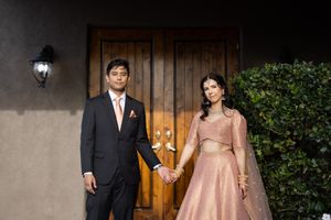 Elegant couple holds hands posing in front of wooden doors, wearing a navy suit and pink lehenga.