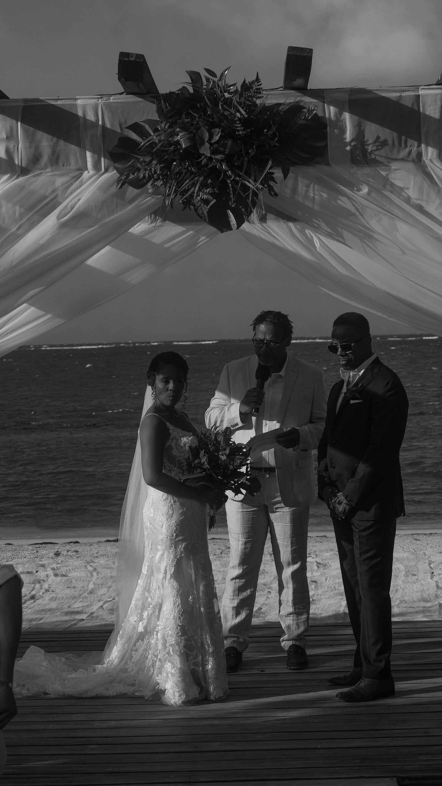 Black and white beach wedding ceremony with bride in lace gown, groom in suit, and officiant under floral arch by the ocean.
