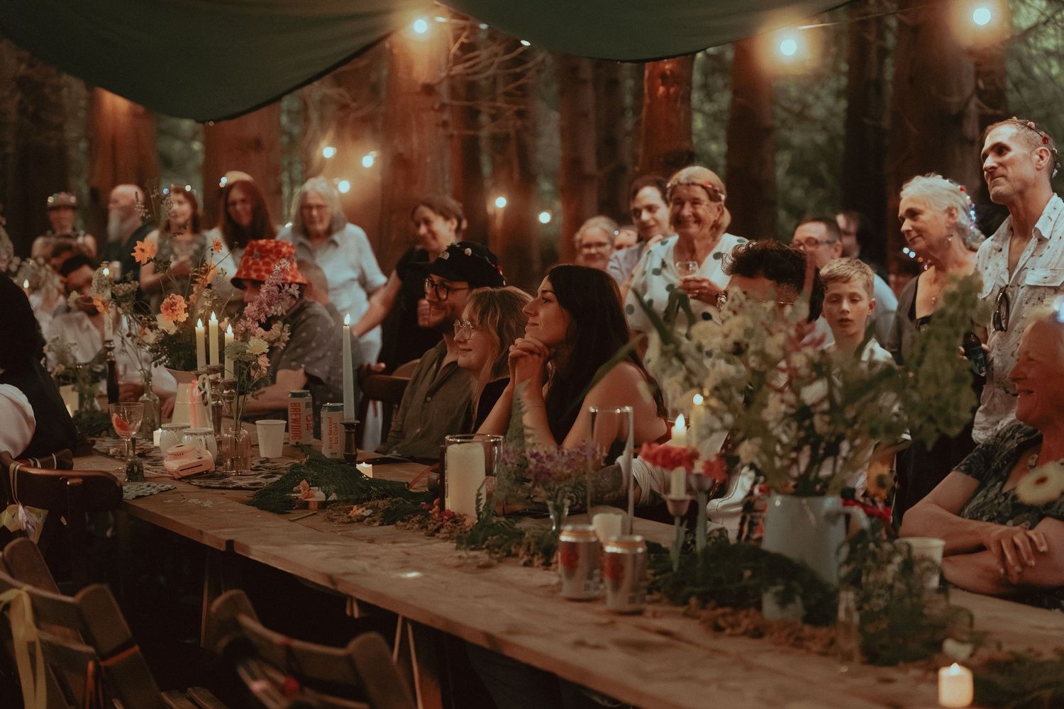 Guests gathered at a rustic outdoor dinner party with string lights, candles, and floral centerpieces in a forest setting.