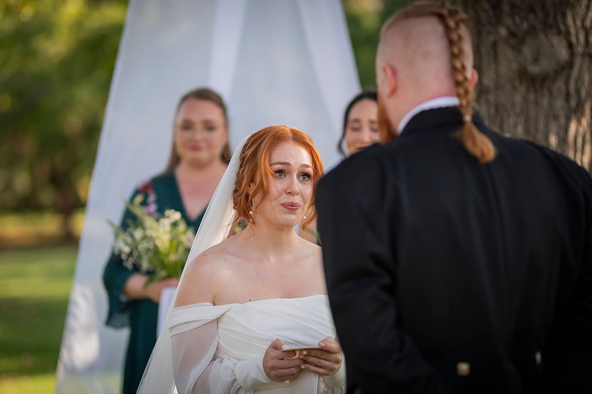 Bride with red hair reads vows at outdoor wedding ceremony, wearing off-shoulder white gown, with bridesmaids standing behind her.