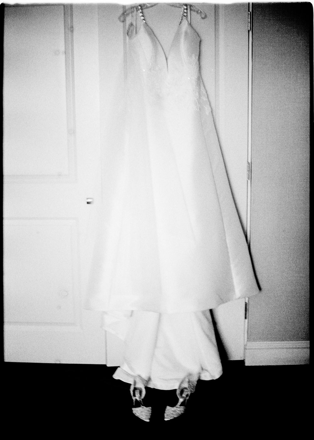 A white wedding gown with lace bodice hangs on a door above bridal heels in a black and white photo.