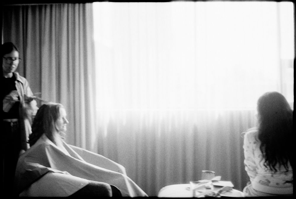Black and white photo of a person getting hair styled in a bright room with curtains, while another person sits nearby.
