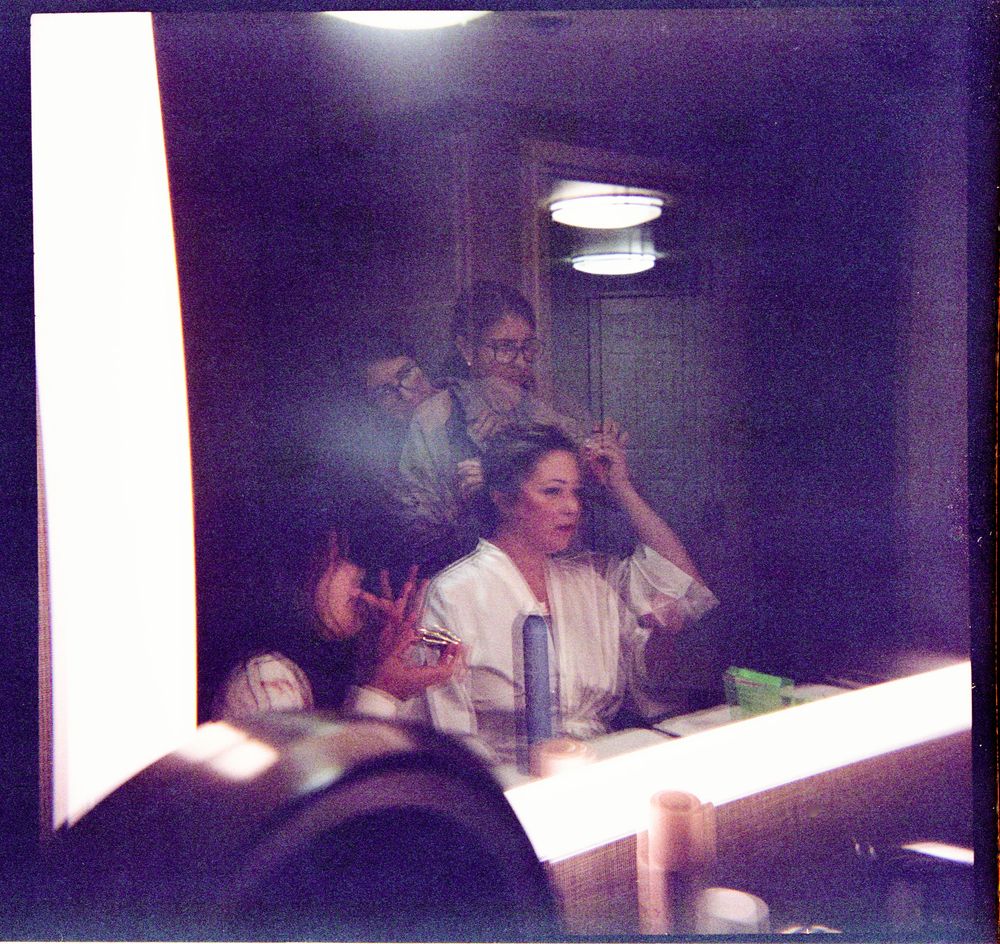 Woman in white robe getting hair styled at a vanity mirror with two hairstylists in a dimly lit room.