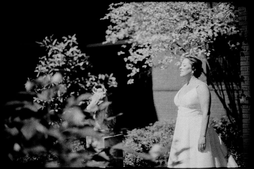 Black and white photo of a bride in a white dress standing outdoors among blooming trees and lush greenery.