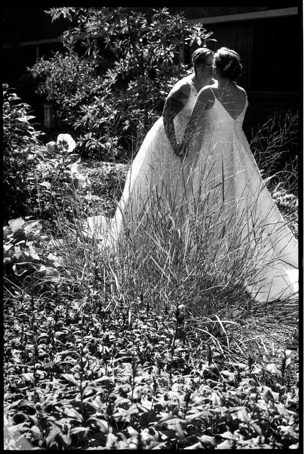 Two brides in white wedding gowns share a romantic moment in a lush garden in black and white photo.