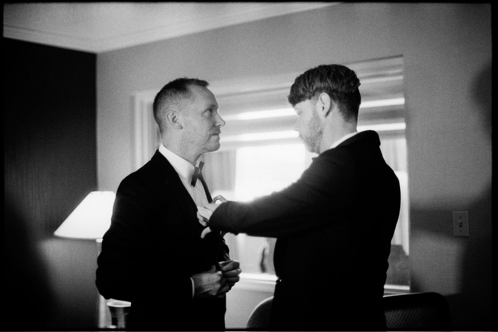 Black and white photo of two men in suits, one adjusting the other's tie before a formal event.