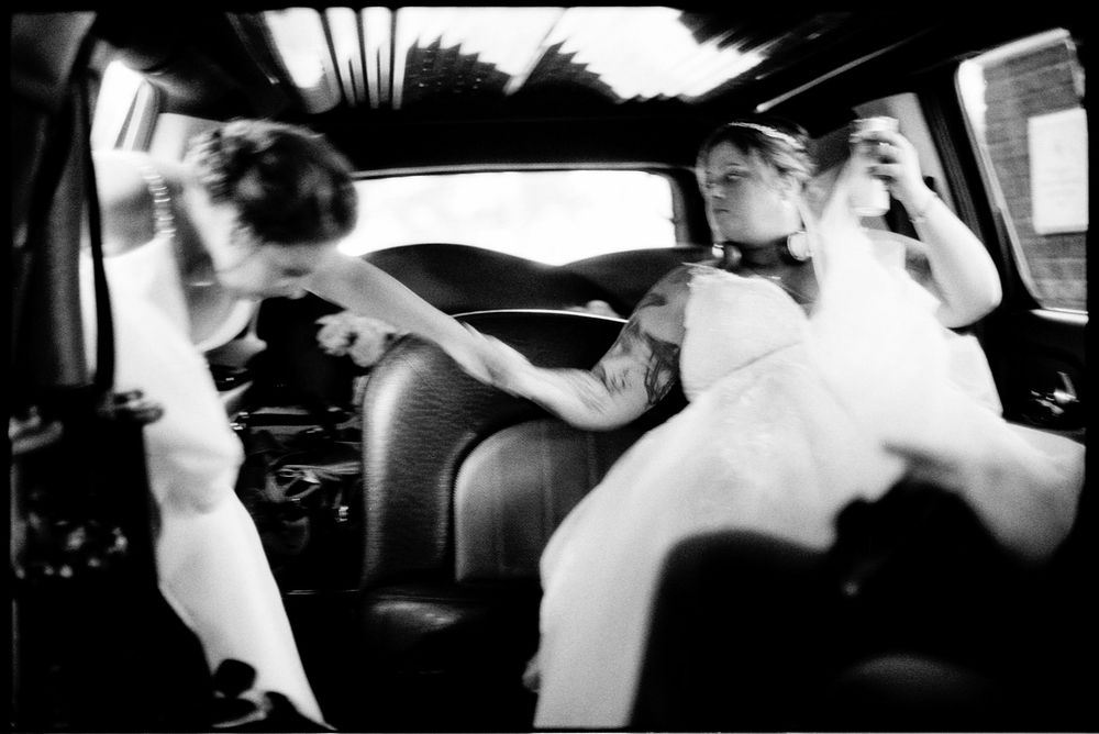 Bride and guests laugh together inside a vintage limousine in a candid black and white wedding photo.