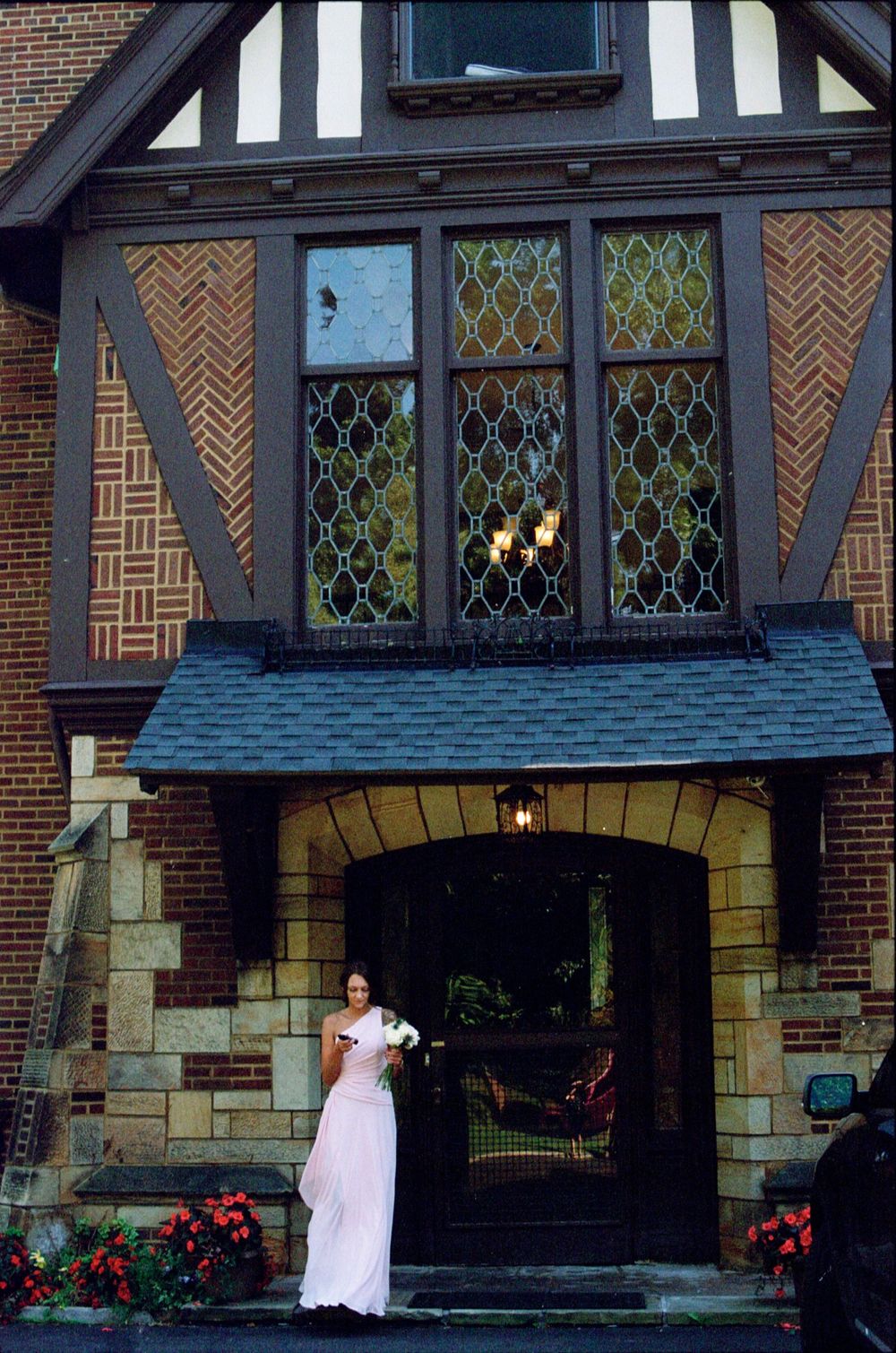 Bridesmaid in pink gown holding white bouquet standing before a Tudor-style brick building.