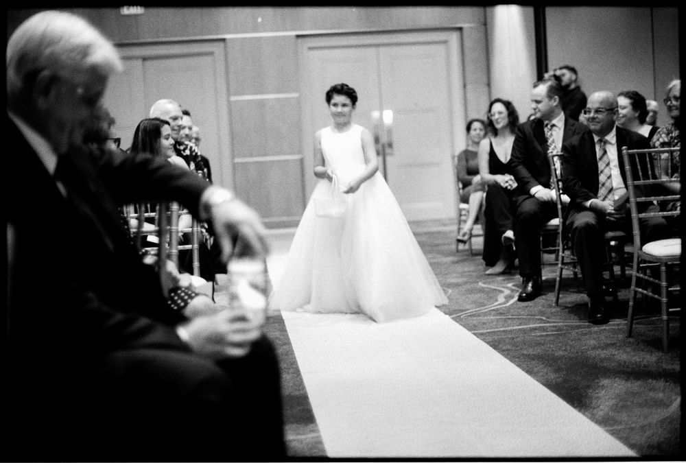 Bride walks down the aisle in a sleek white gown during a formal wedding ceremony in black and white.