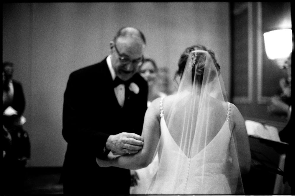 Father escorts bride down the aisle in an emotional black and white wedding ceremony photo.