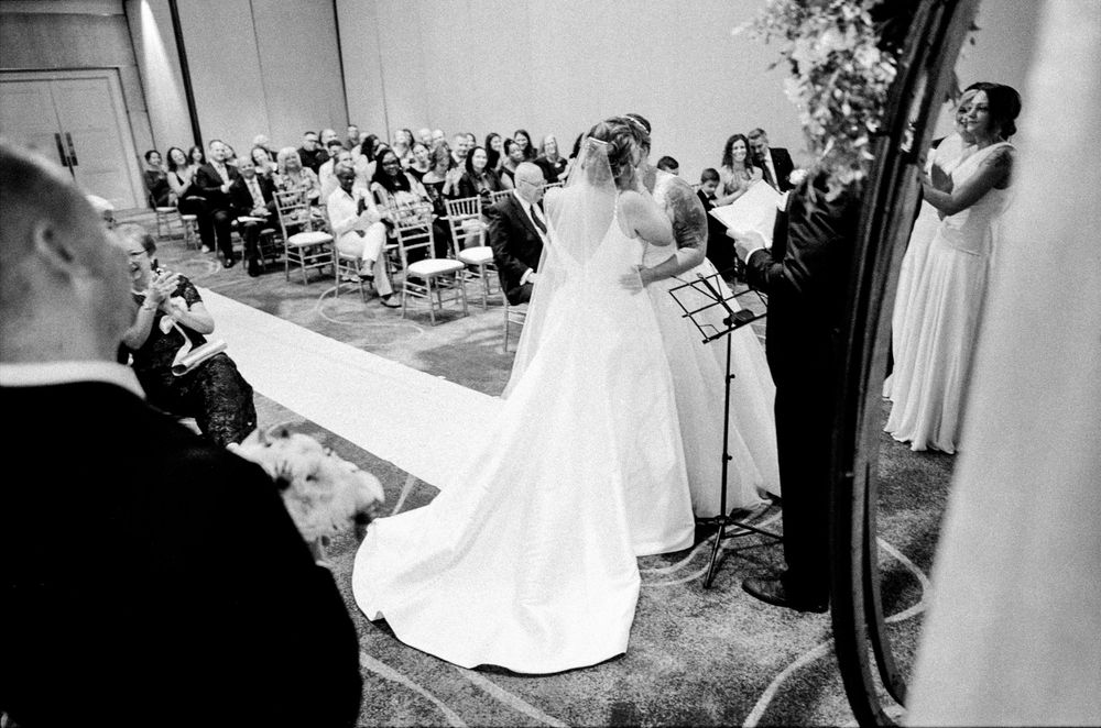Black and white wedding ceremony photo showing bride in flowing gown at altar with guests seated in elegant venue.