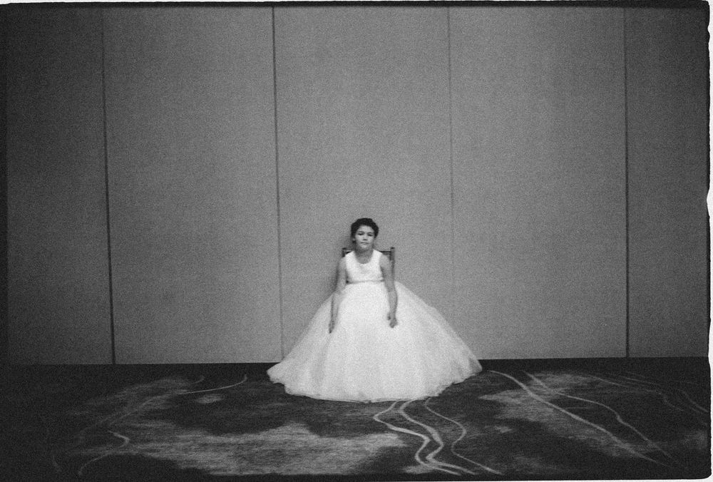 Black and white photo of a bride in a voluminous white ball gown standing alone in a large empty ballroom.