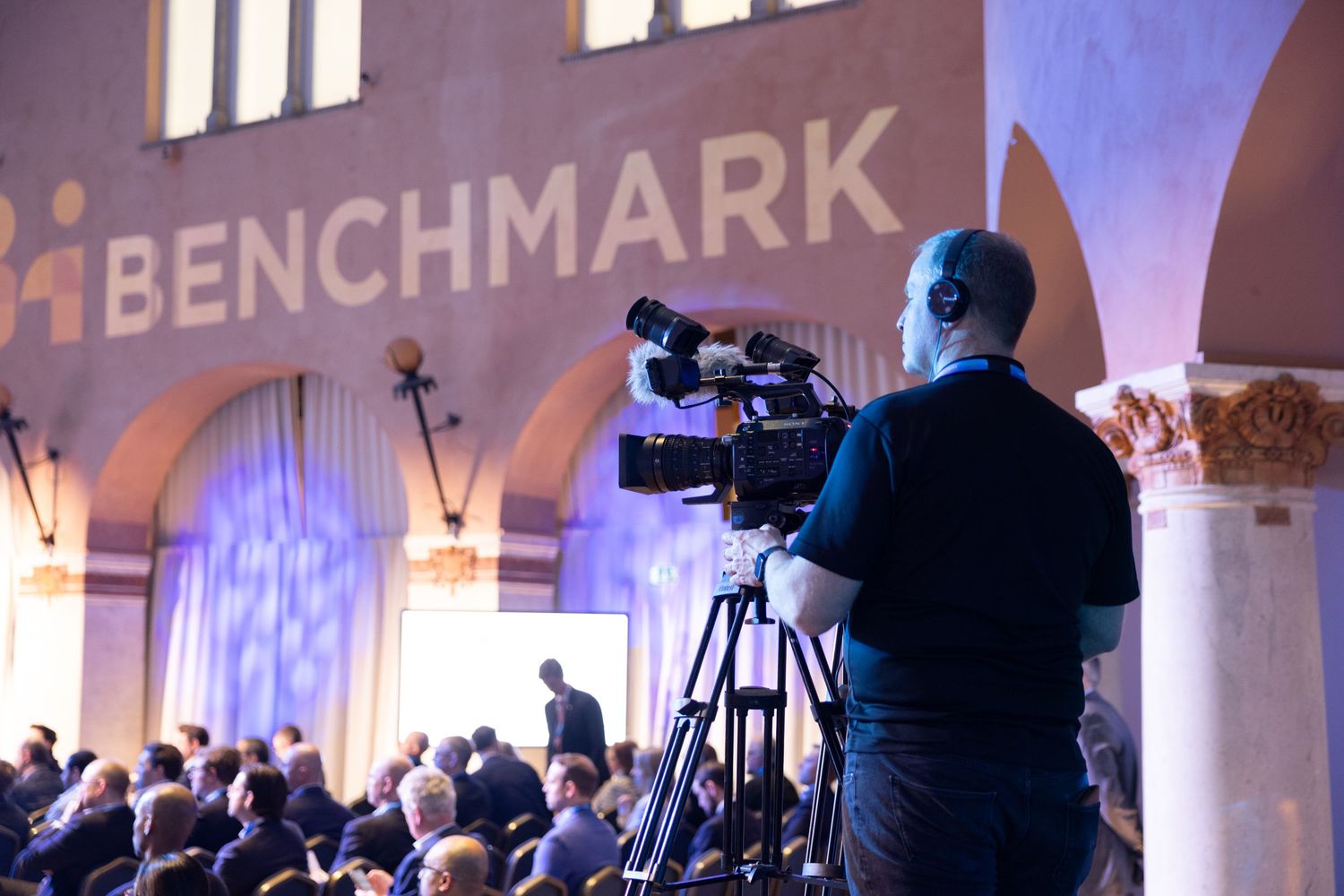 Videographer filming a large Benchmark-branded conference event from behind the audience.