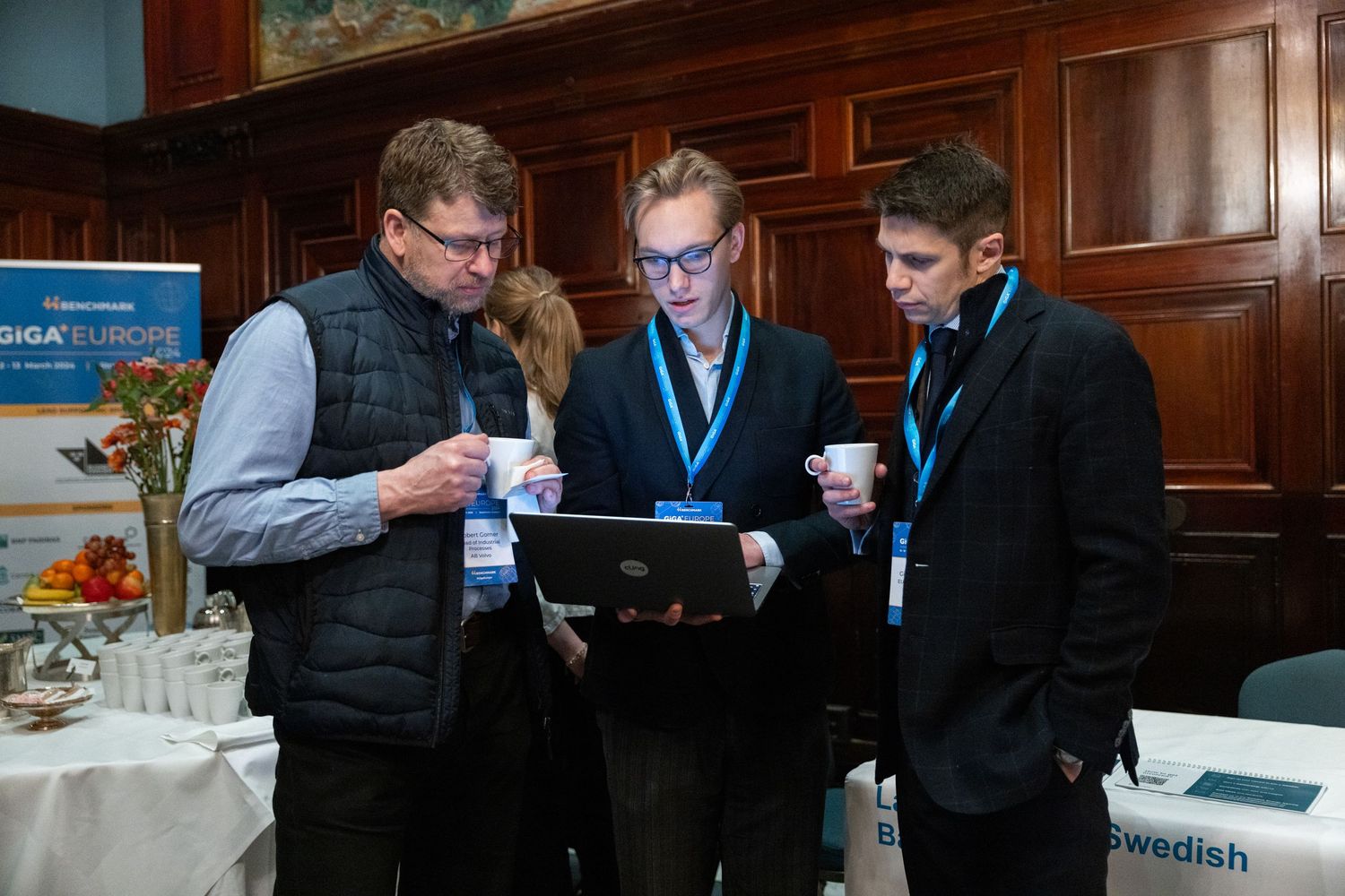 Three men reviewing content on a laptop at the GiGA Europe conference networking area.