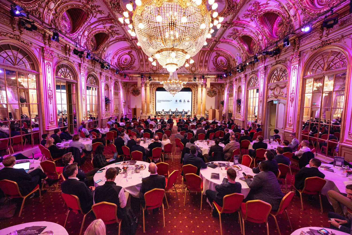 Elegant ballroom conference with round tables and attendees seated beneath ornate chandeliers and pink lighting at Grand Hotels Spegelsalen