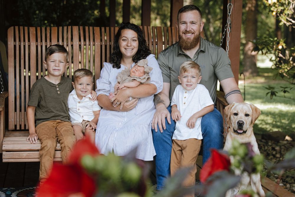 Family portrait on porch swing with three boys, newborn baby, yellow Labrador, and parents smiling outdoors.