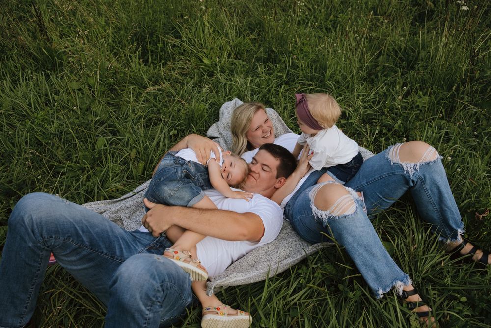 Family of four laughing and cuddling together in lush green grass, wearing casual jeans outfits during outdoor photo session.
