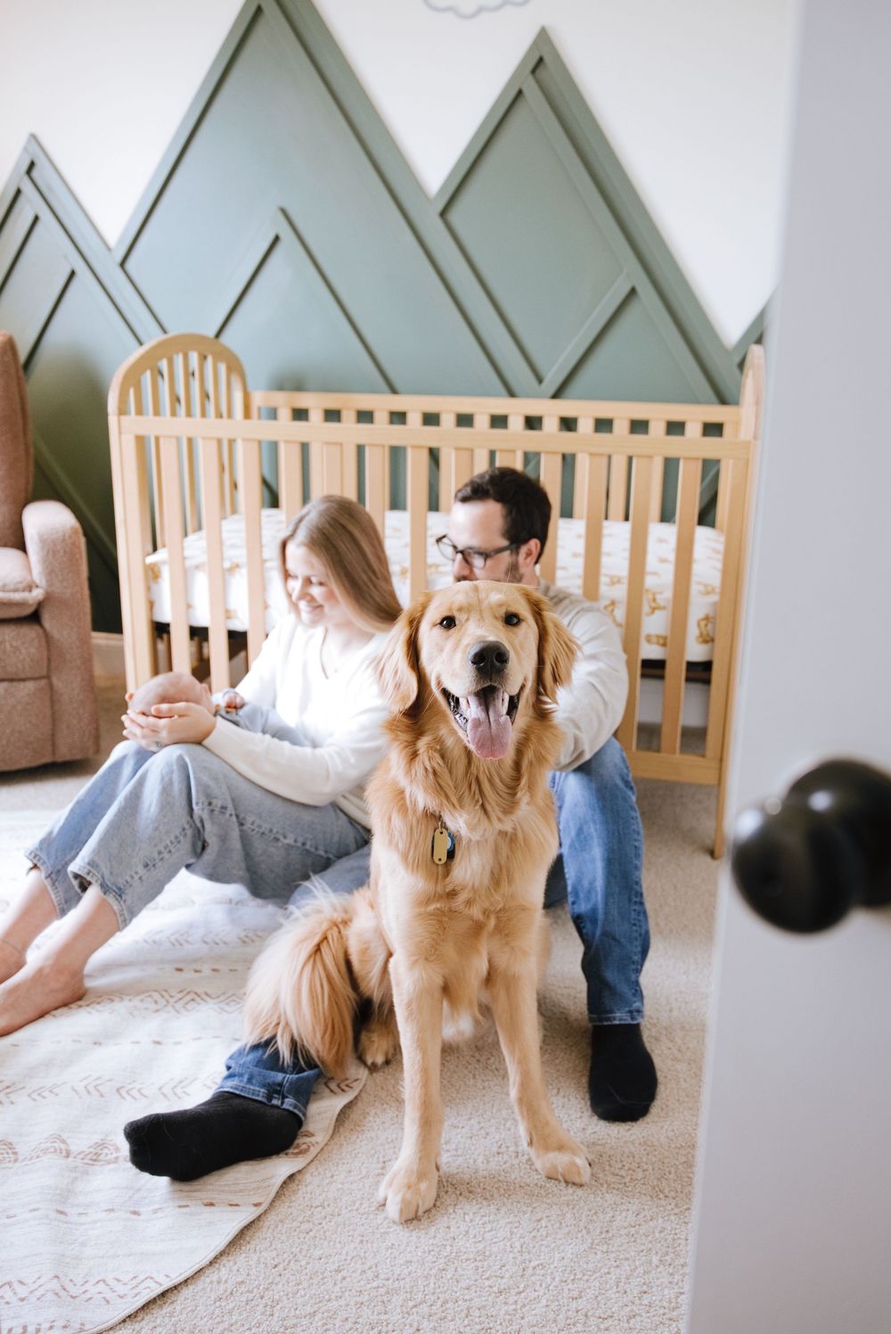 Couple sits on nursery floor with golden retriever dog in front of wooden crib with mountain wall mural.