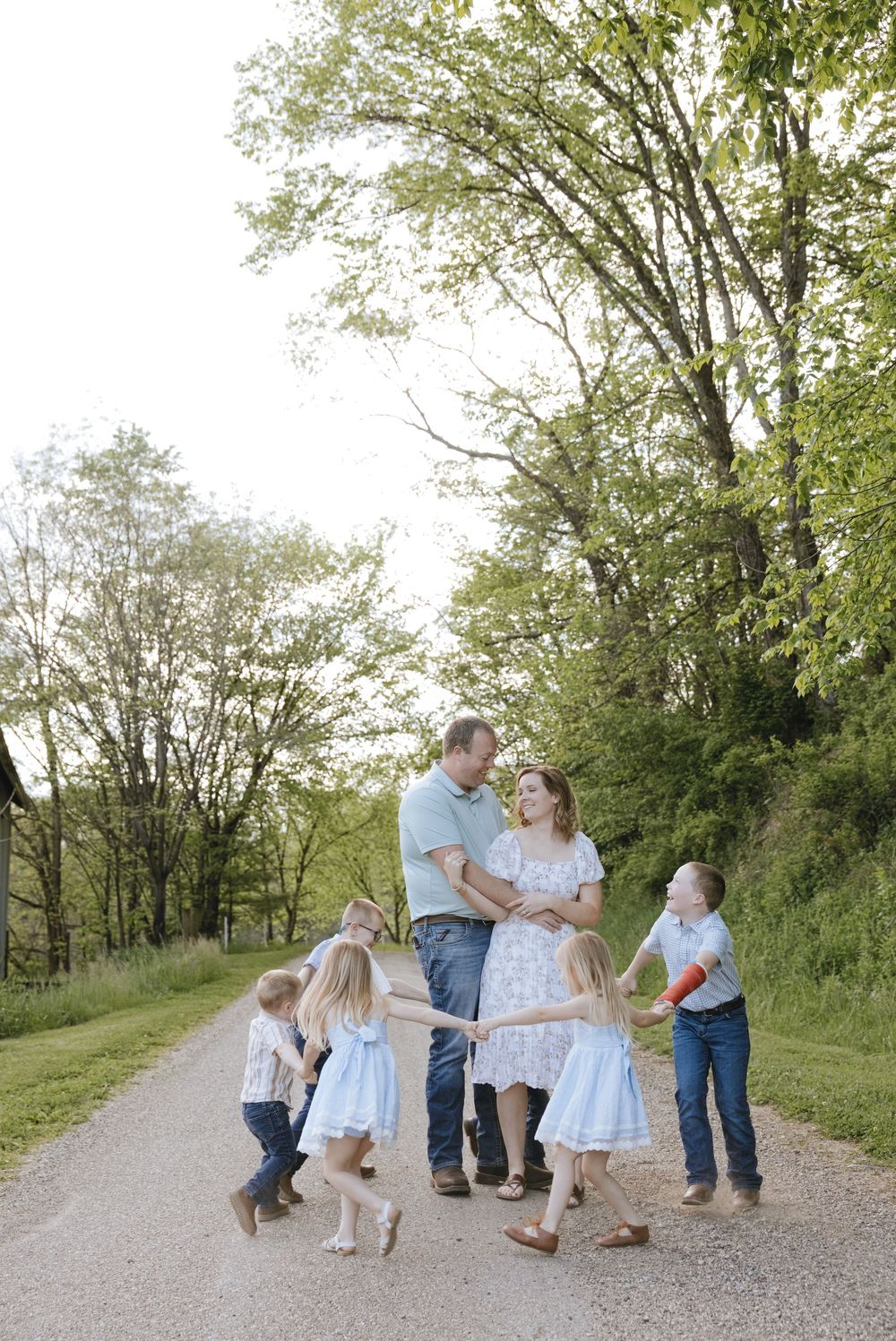Family of six poses on a scenic country road surrounded by lush green trees during an outdoor photo session.