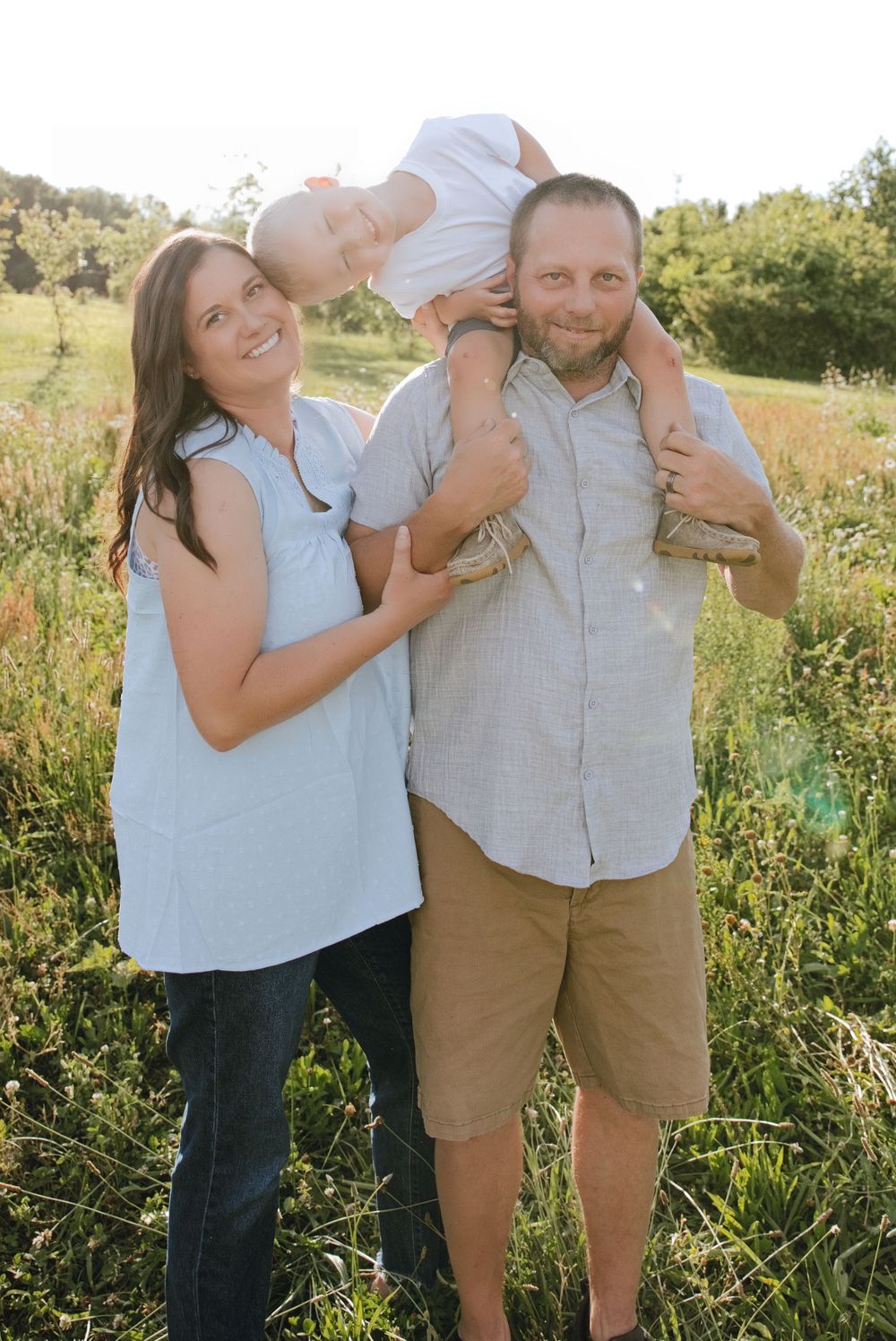 A happy family poses outdoors in a sunny field, with dad holding a baby on his shoulders while mom smiles beside them.