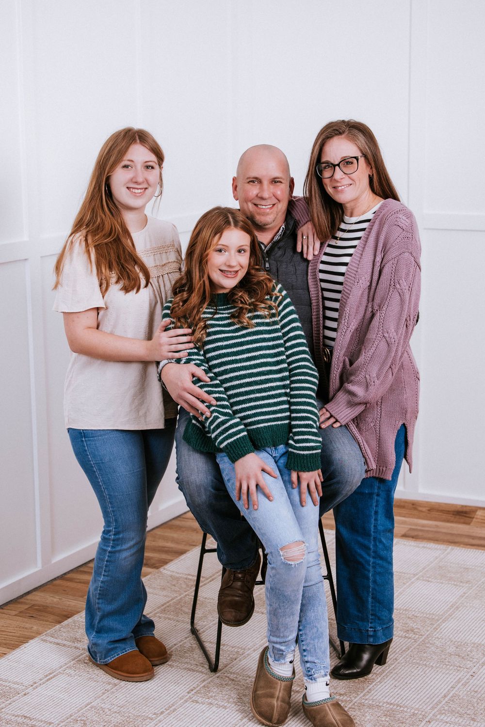 A bald man sits on a chair surrounded by three smiling girls in casual attire, posing for a family portrait indoors.