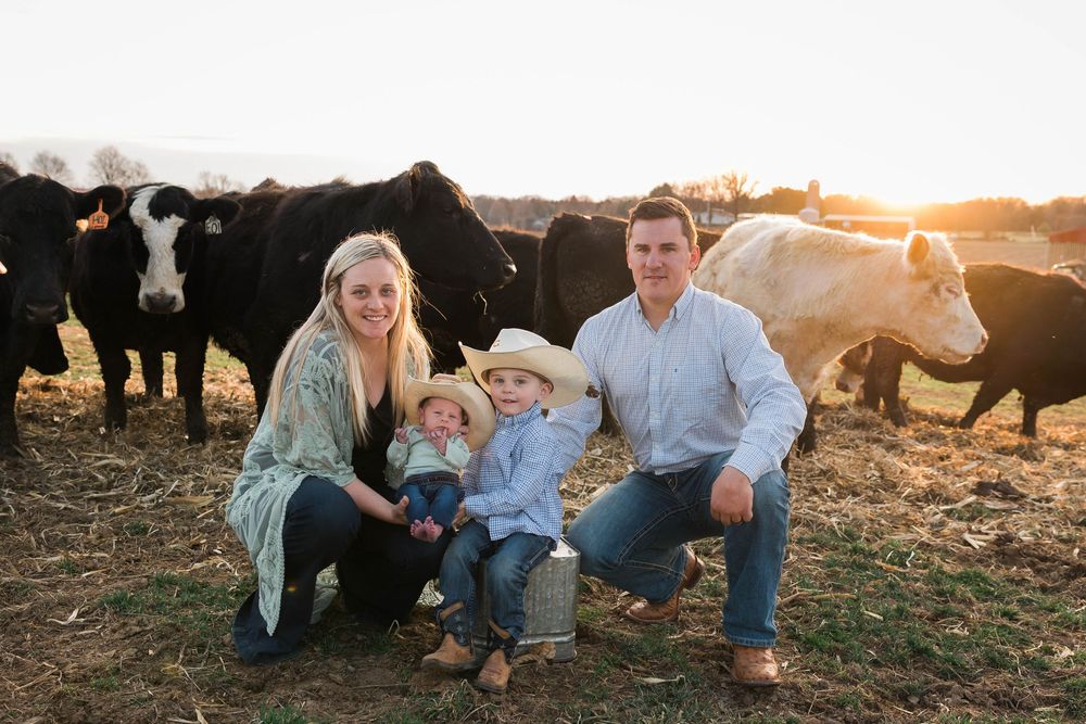 Family of four poses with cattle herd at golden hour on a farm, parents and two young children in cowboy hats.