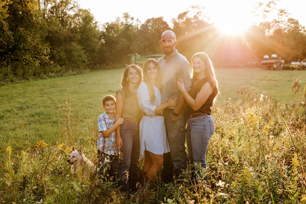 Family of six posing together in a sunlit field during golden hour, surrounded by wildflowers and lush green trees.