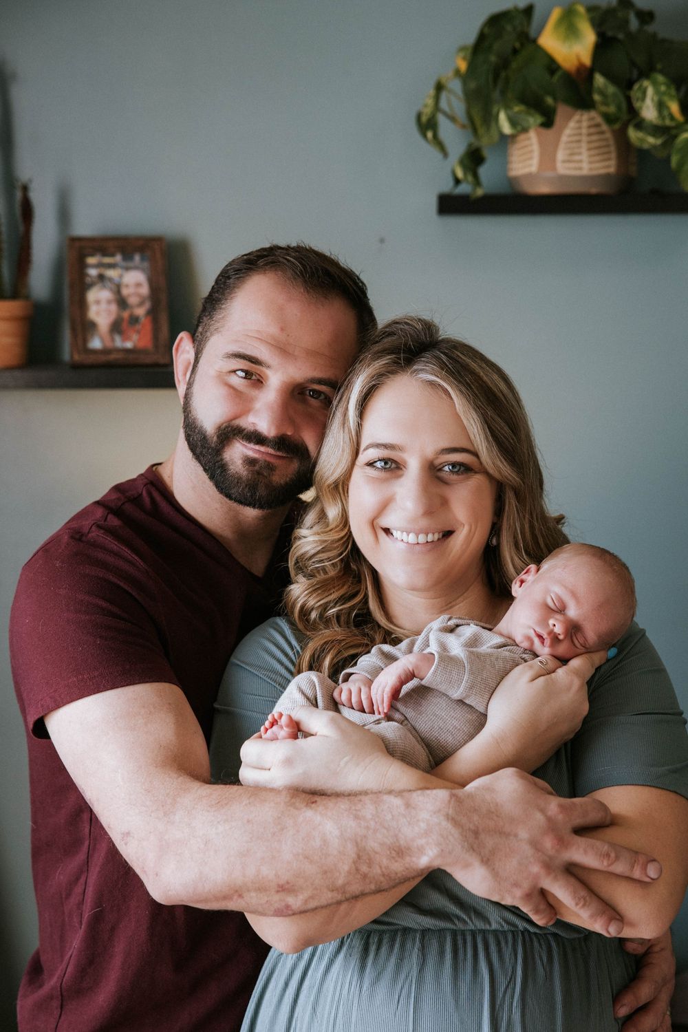 Happy couple smiling and holding their newborn baby in a cozy home setting with plants and decor on shelves.