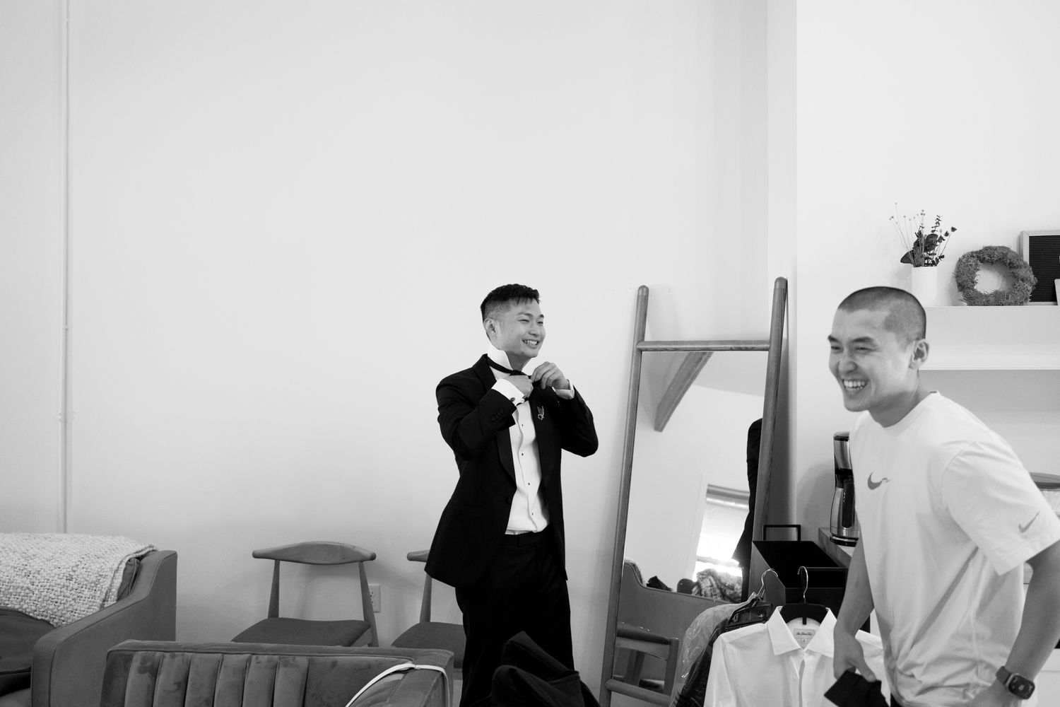 Two men laughing and smiling in a bright room, one dressed in a black suit during wedding preparations.