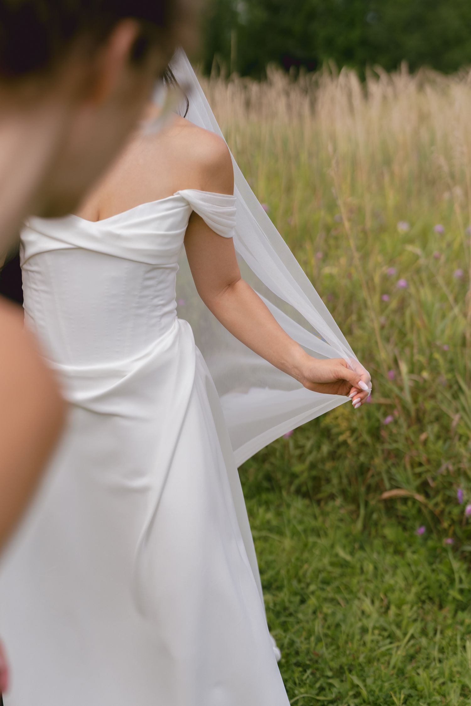 Close-up of bride in off-shoulder white wedding gown holding veil in green meadow field.