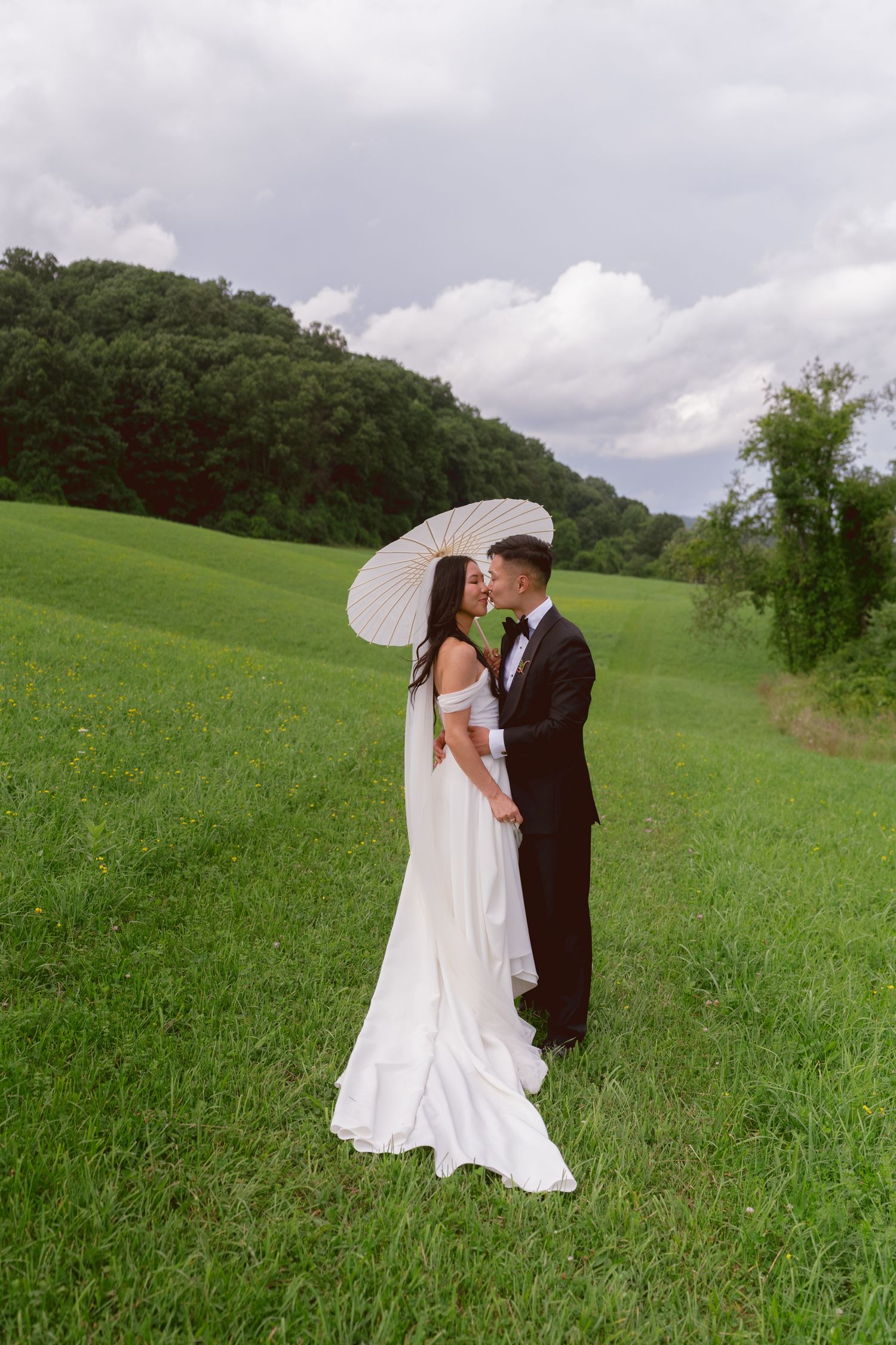 Bride and groom share a kiss in a lush green field, bride in white gown holding a parasol umbrella.