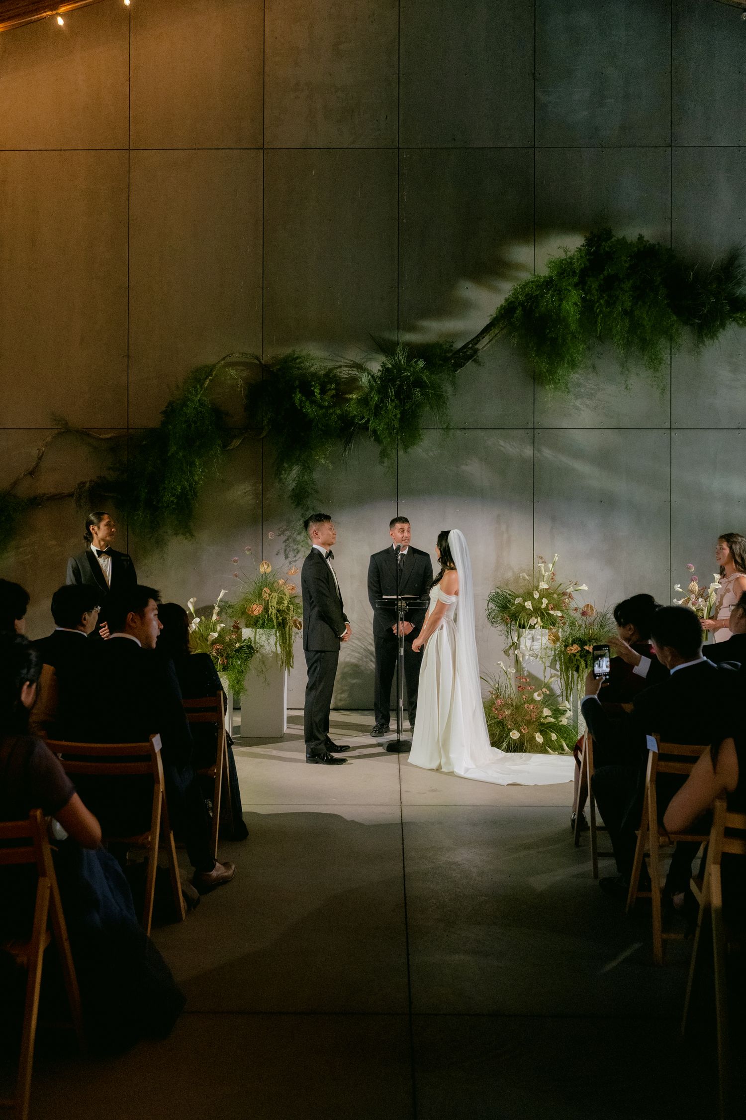 Wedding ceremony in a modern venue with stone walls, greenery garland, and guests seated in wooden chairs.