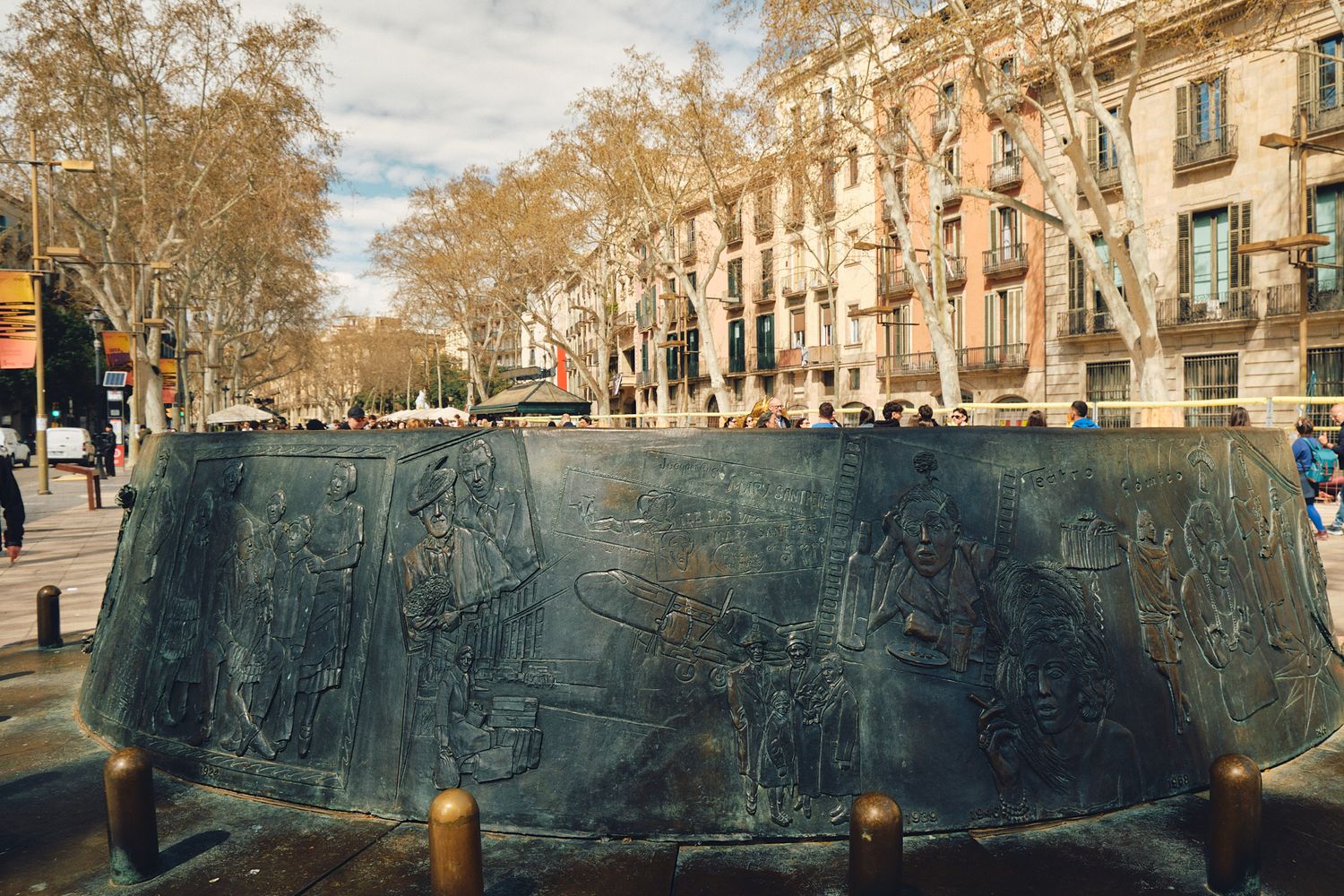 Bronze sculptural monument with relief carvings on Las Ramblas boulevard in Barcelona, Spain, surrounded by trees.