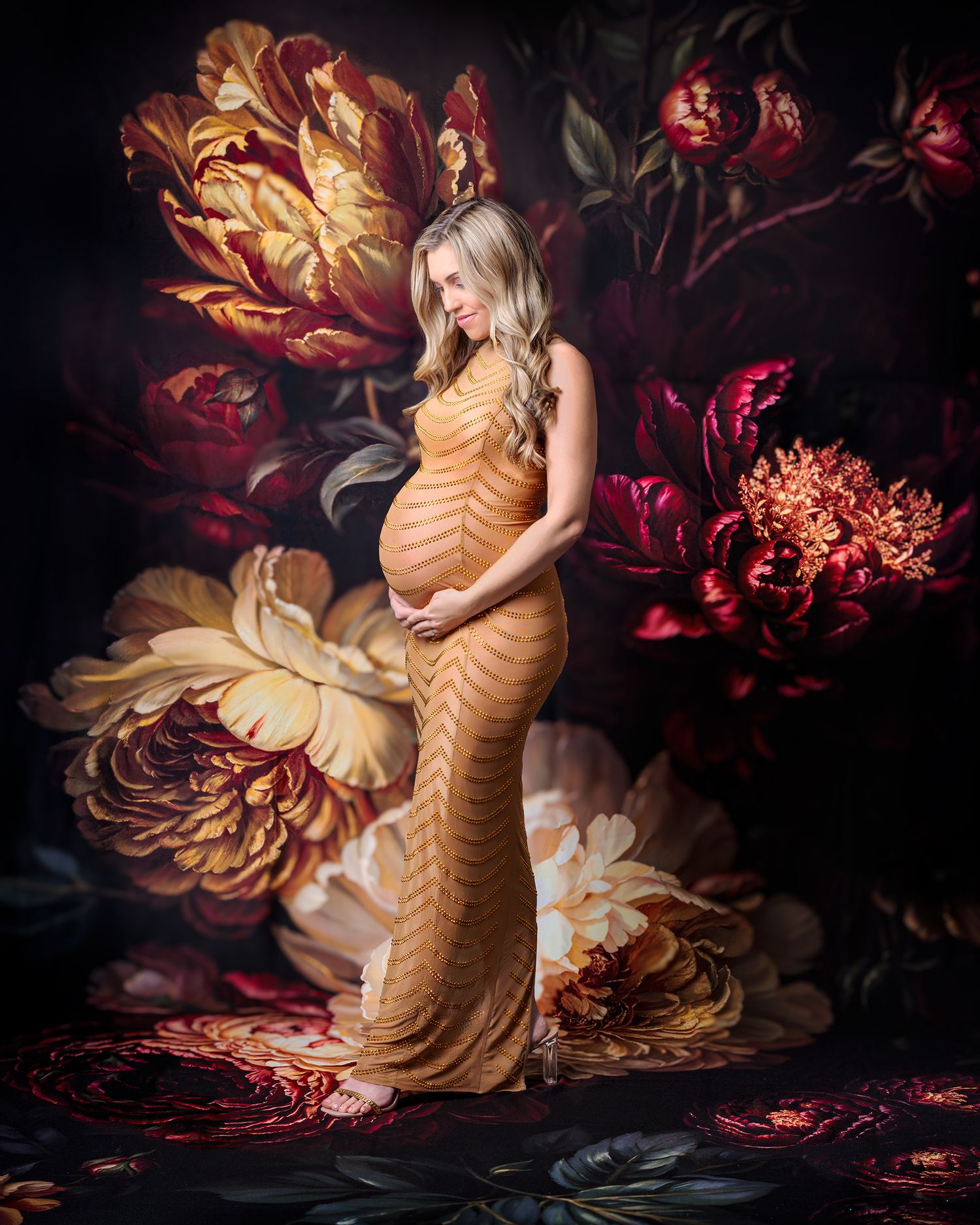Elegant maternity portrait of a woman in a figure hugging gown with gold detail lovingly holding her pregnant belly against moody floral set