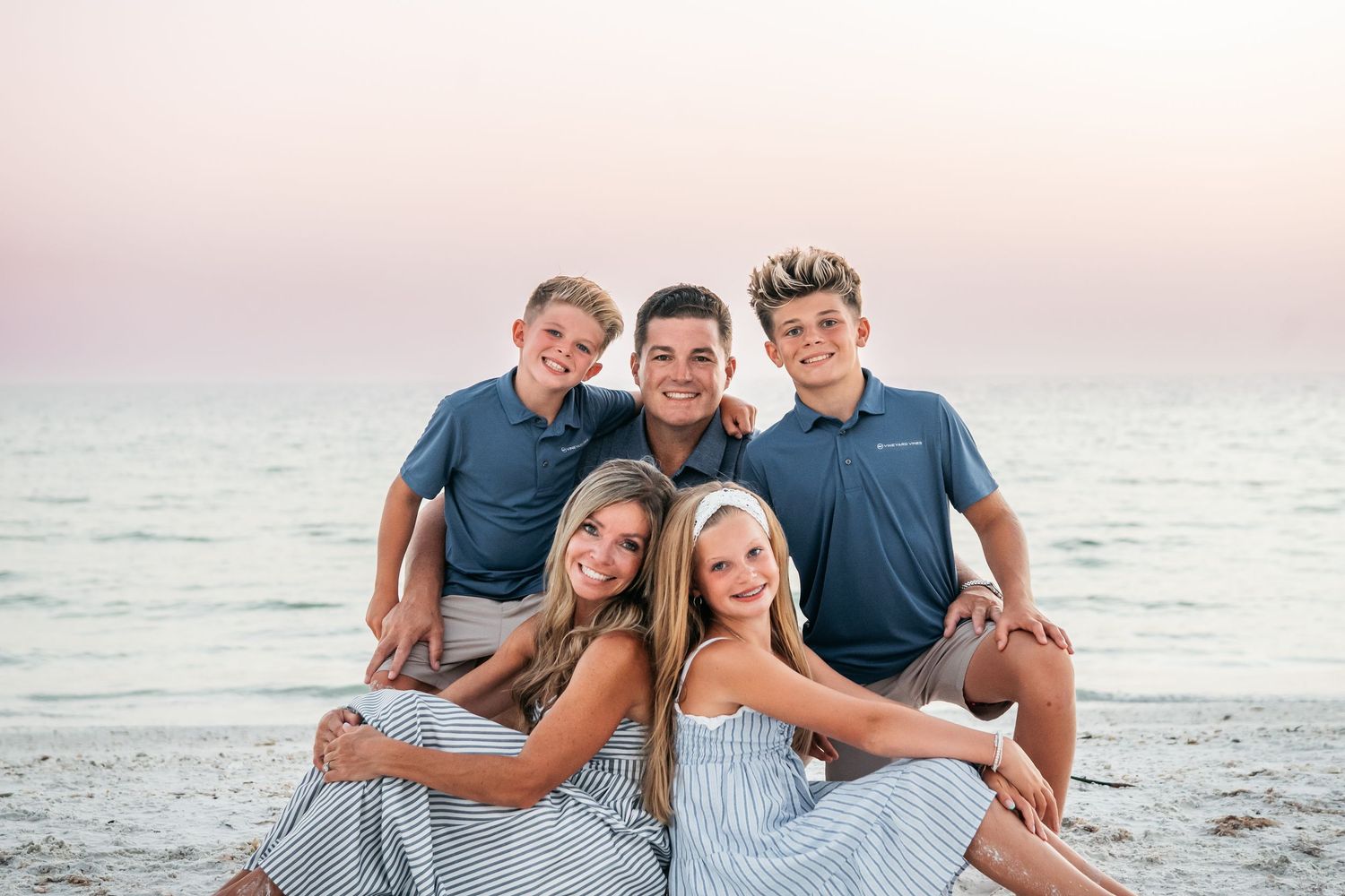 Family of five smiling together on a beach at sunset, wearing coordinating blue outfits.