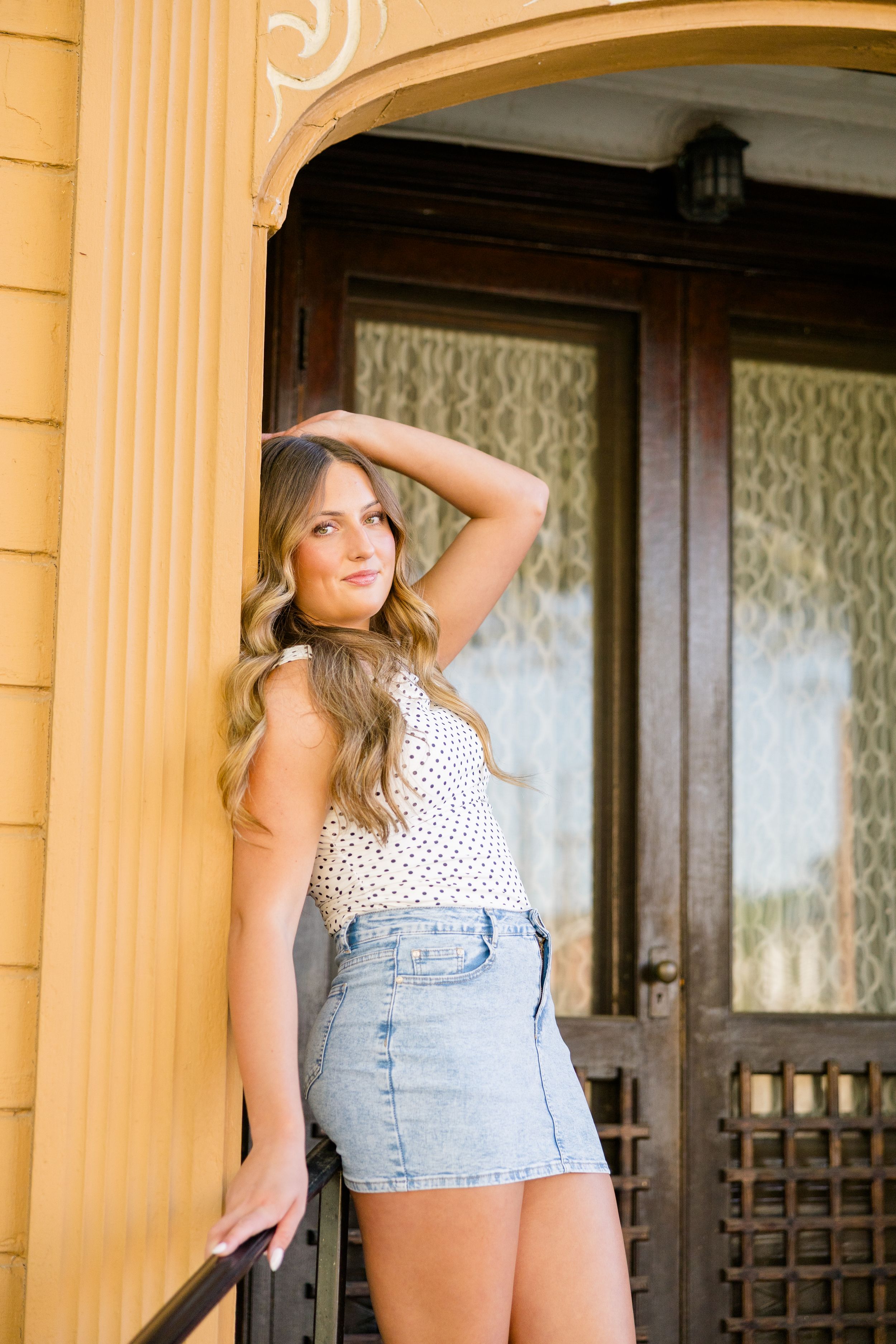 Young woman in denim skirt and polka dot top posing by ornate wooden door of yellow building.