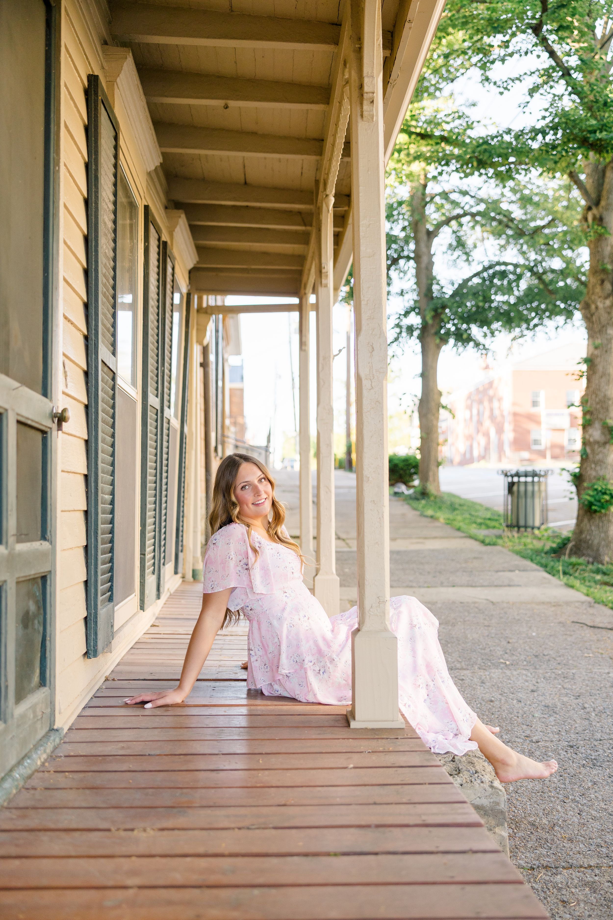 Woman in pink floral maxi dress sitting barefoot on wooden porch of historic building, smiling outdoors.