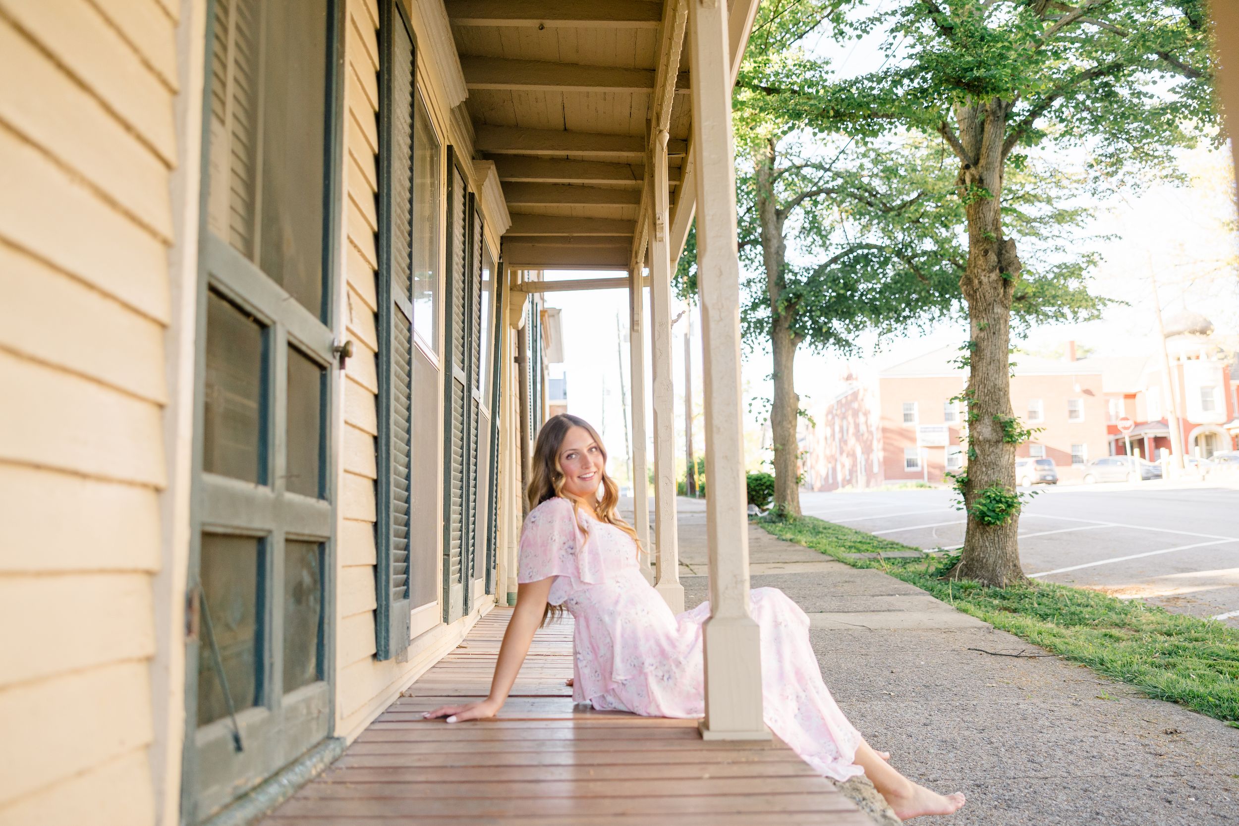 Young woman in pink floral dress sitting on wooden porch of historic building, smiling outdoors.