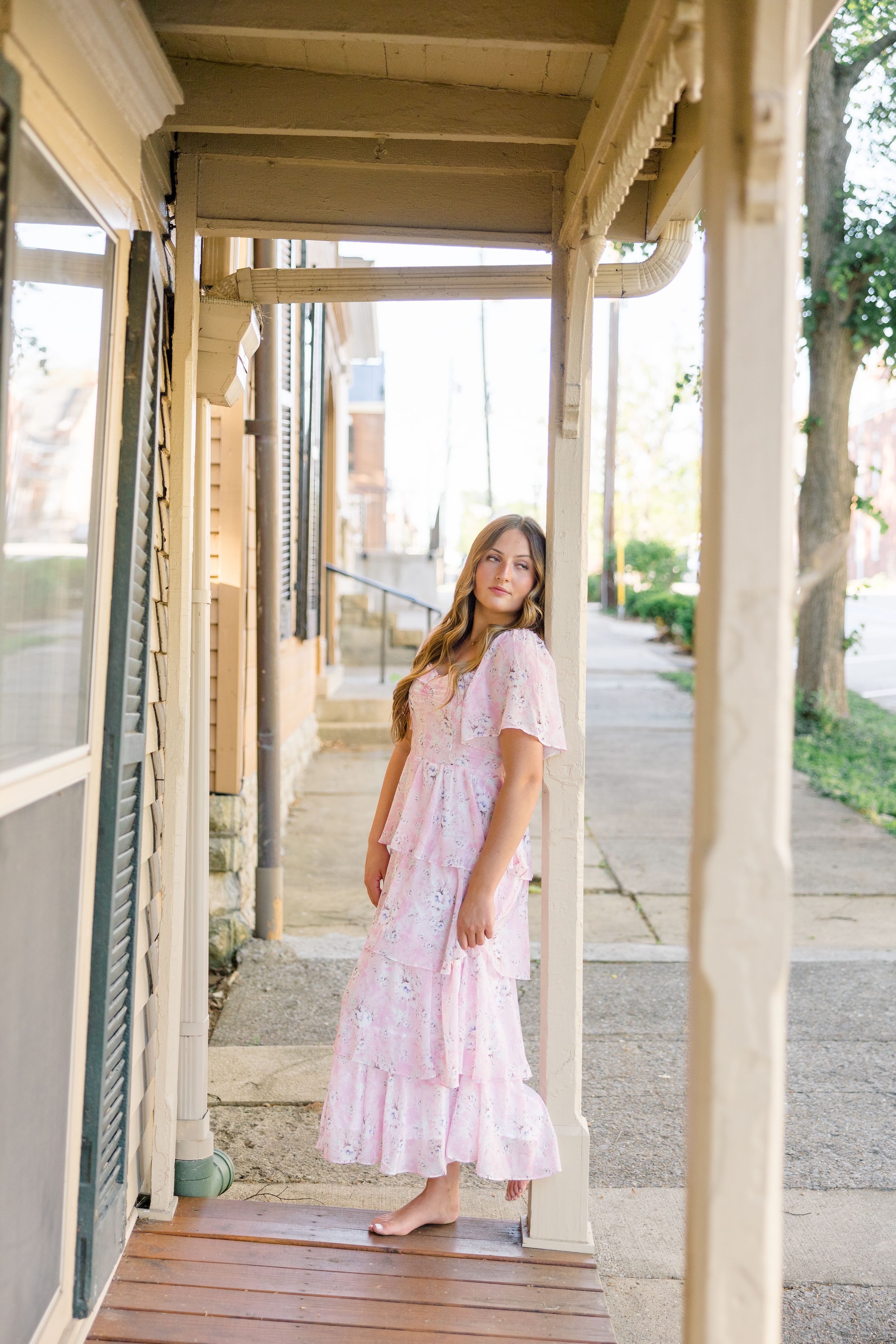 Young girl in a pink floral maxi dress poses on a wooden porch of a quaint small-town storefront.