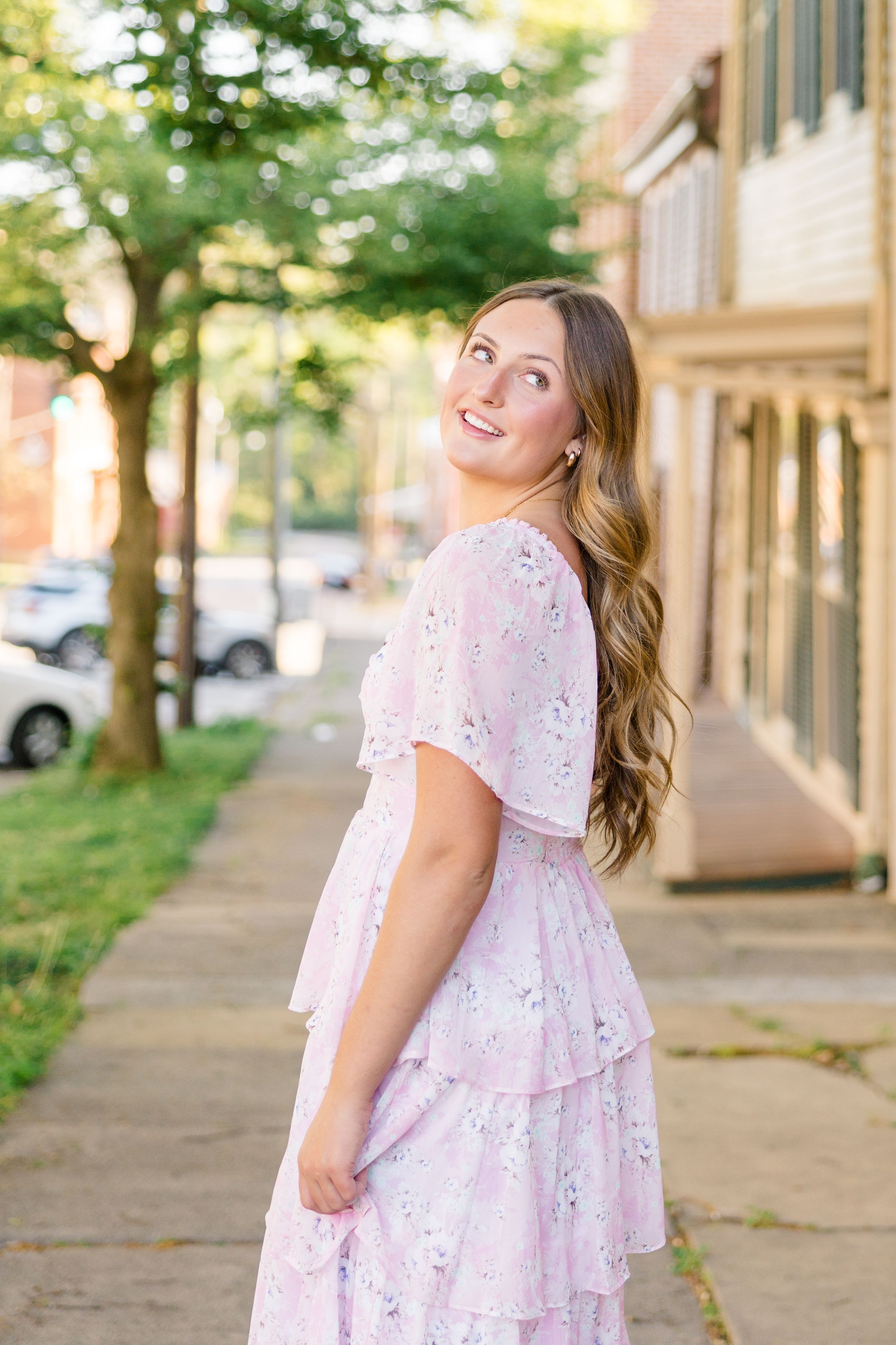 Young woman in pink floral tiered dress smiling over shoulder on tree-lined sidewalk in golden hour light.