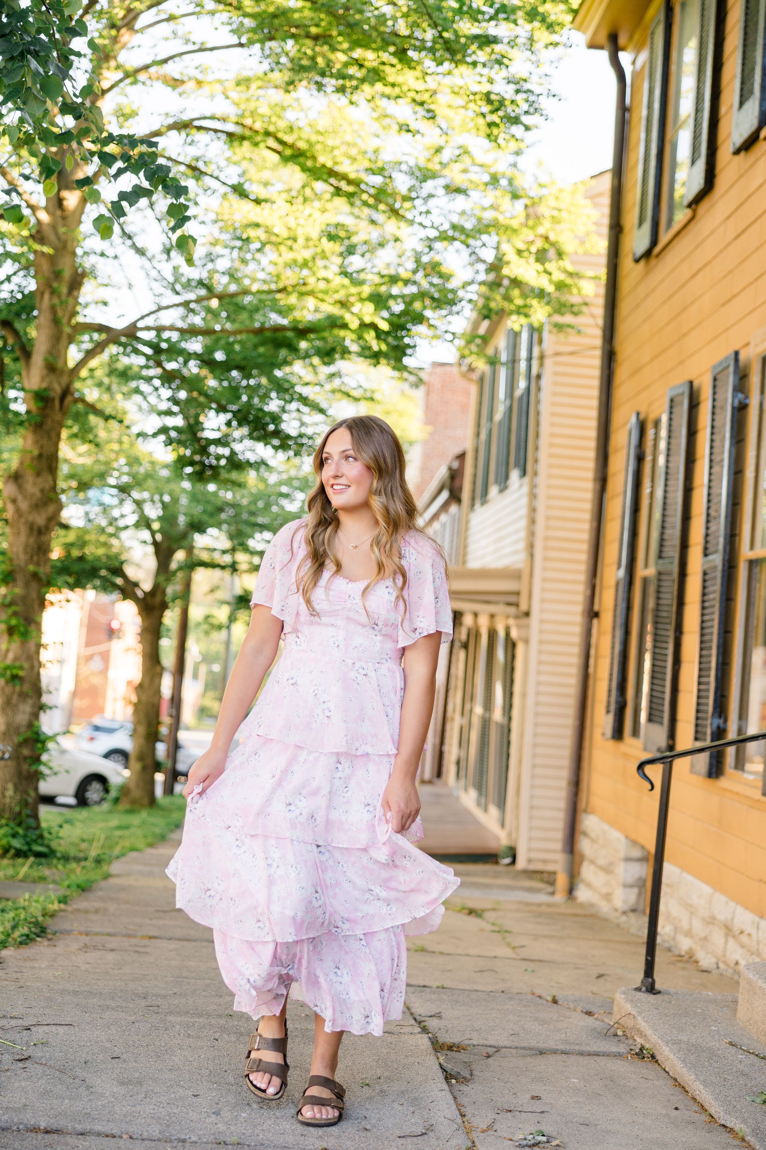 Young woman in a pink floral tiered maxi dress walking on a sidewalk beside a yellow building with lush green trees.