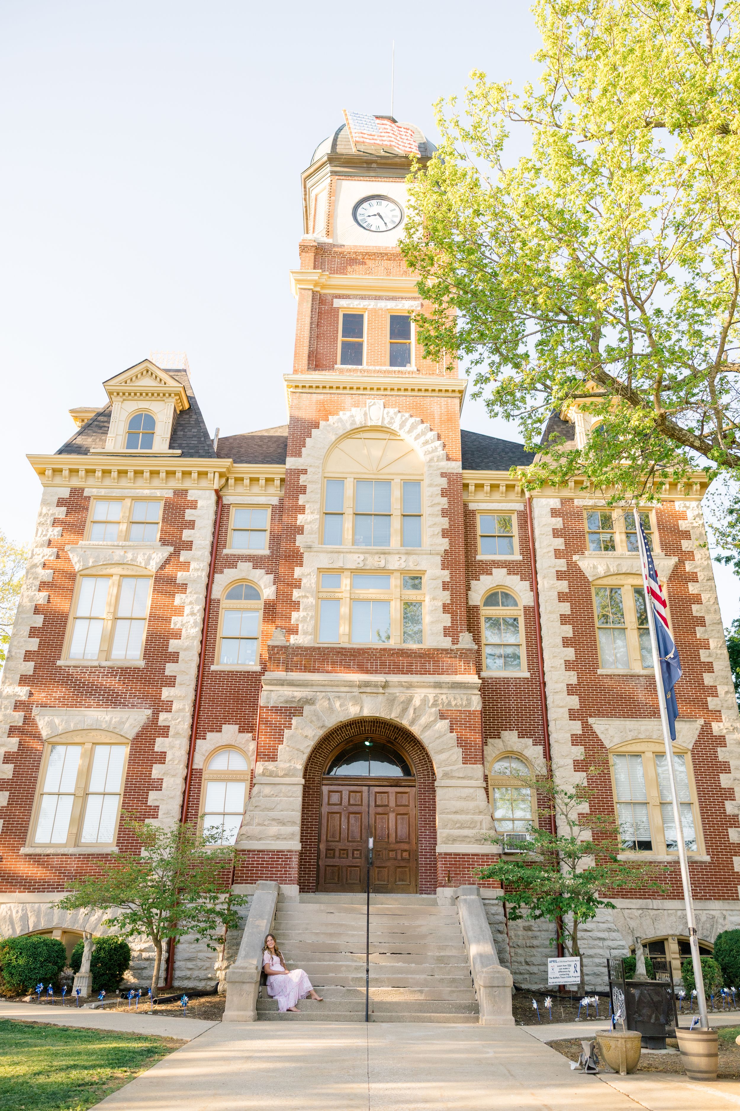 Woman in white dress sitting on steps of historic red brick courthouse with clock tower and American flag.