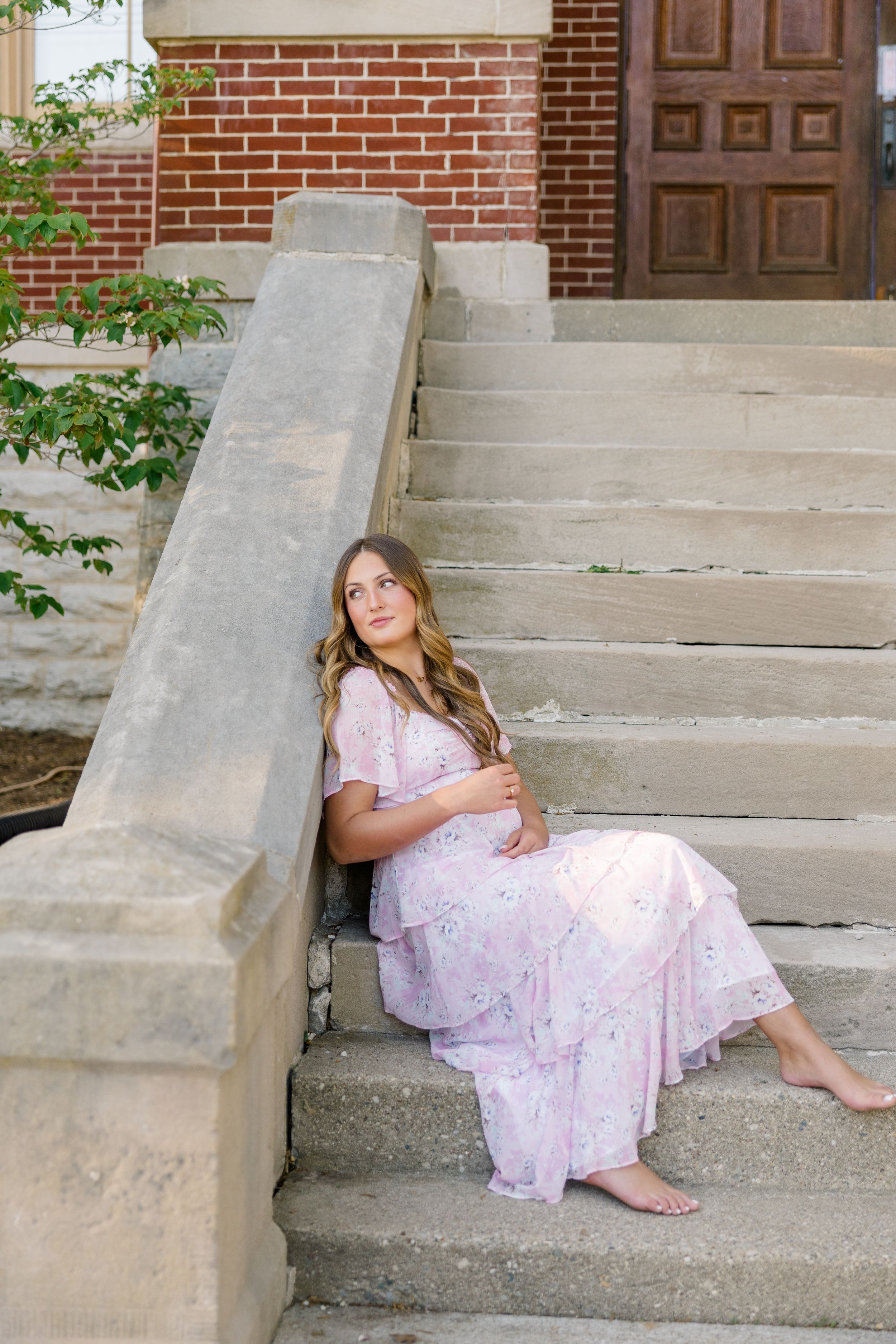 Pregnant woman in pink floral dress sitting barefoot on stone steps outside brick building.
