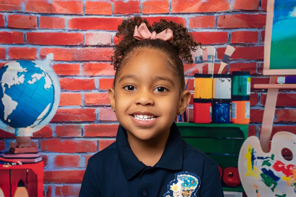 Cheerful young girl in navy uniform smiles with pink bow in hair against brick school backdrop.