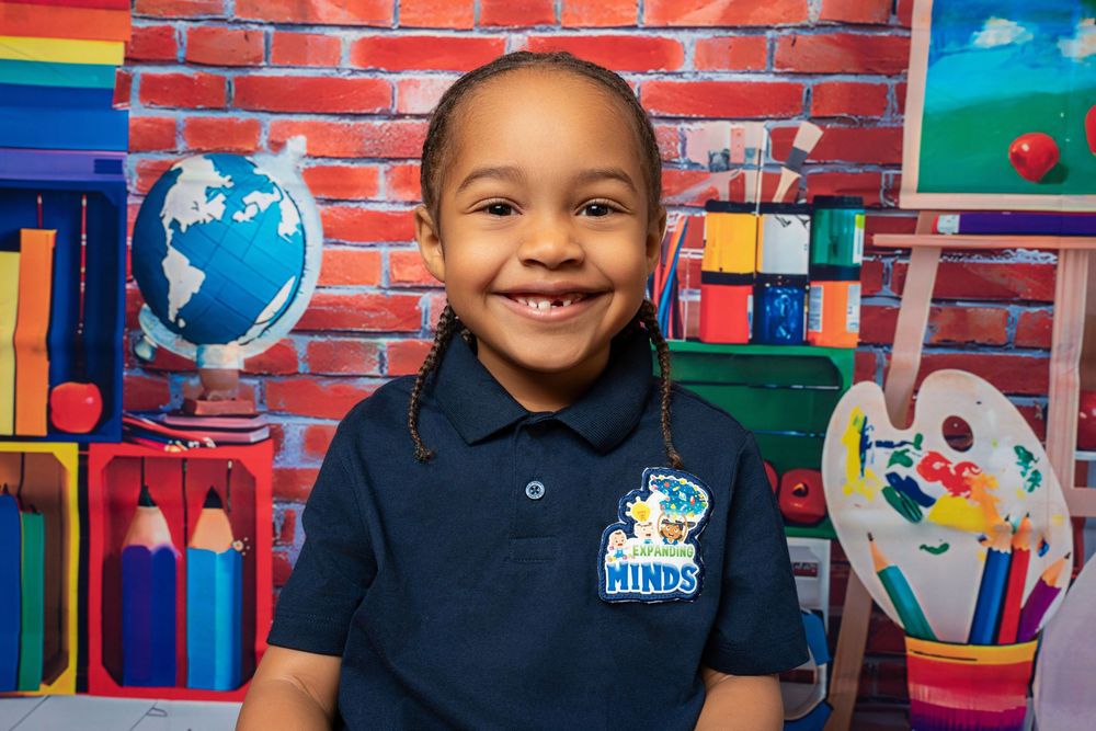 Bright-eyed child in navy Hinds school polo grins before colorful brick classroom backdrop.