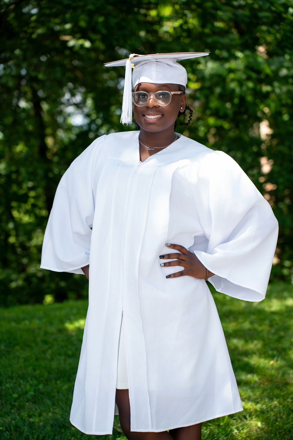 Graduate in white cap and gown standing outdoors with hand on hip among green trees.