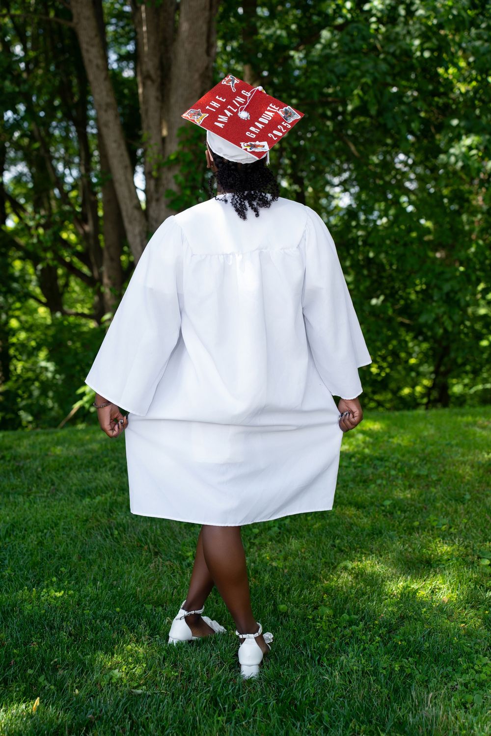 Graduate in white gown with decorated red cap walking away in lush green outdoor setting.