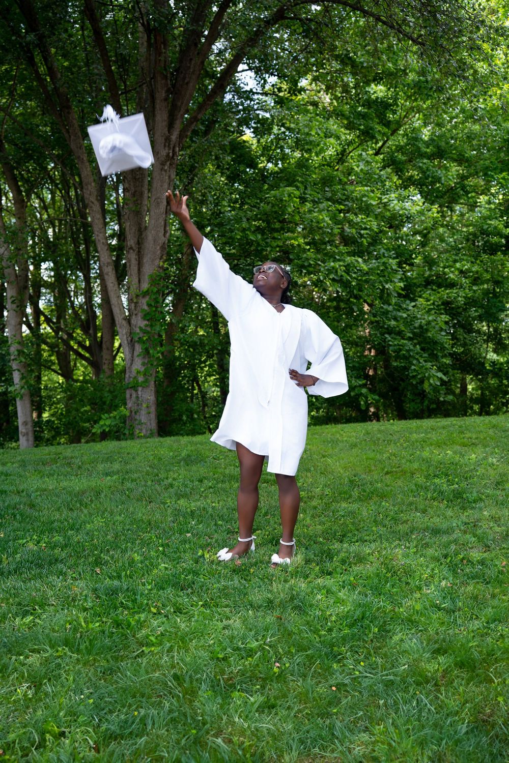 A joyful graduate in white cap and gown tosses her cap in the air outdoors on green grass.